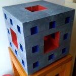 Opengewerkte kubus Beeld bestaat uit 8 delen, ingezaagd. Totale maat 36x36x36 cm, Belgisch hardsteen. Idee achter het beeld is dat door de inzagingen er een beeld ontstaat met minimale inhoud en maximaal oppervlak. Het binnenwerk van het beeld is in rood en blauw geschilderd.